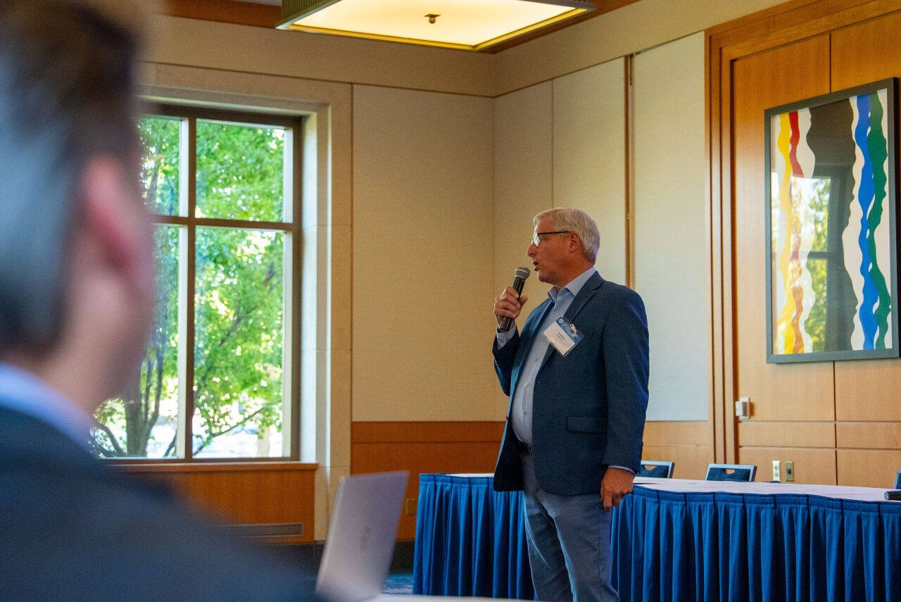 Jonathan Engelsma, professor in the College of Computing, speaks into a microphone while presenting at the Tech Week Grand Rapids session “Connecting Research and Industry to Fuel Tech Growth.” He stands near a window with natural light streaming in.